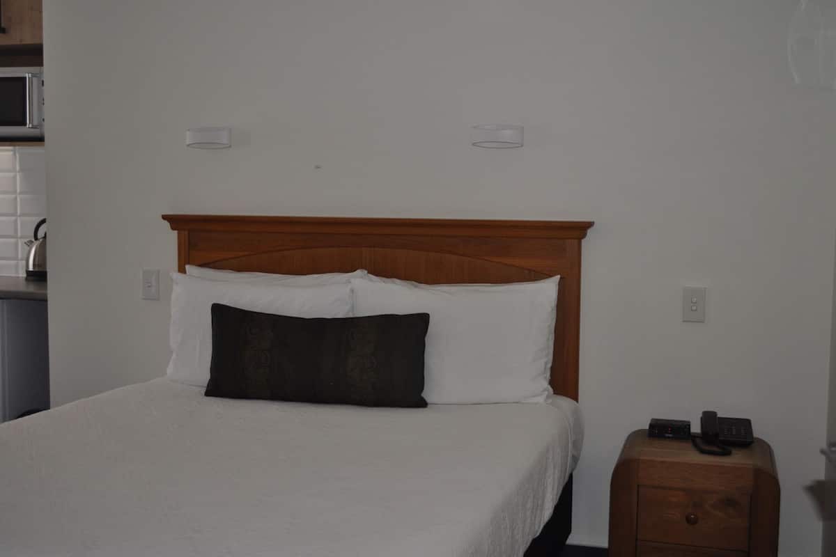Una cama con almohadas blancas y oscuras, cabecera de madera, mesita de noche con un teléfono y un microondas al fondo.