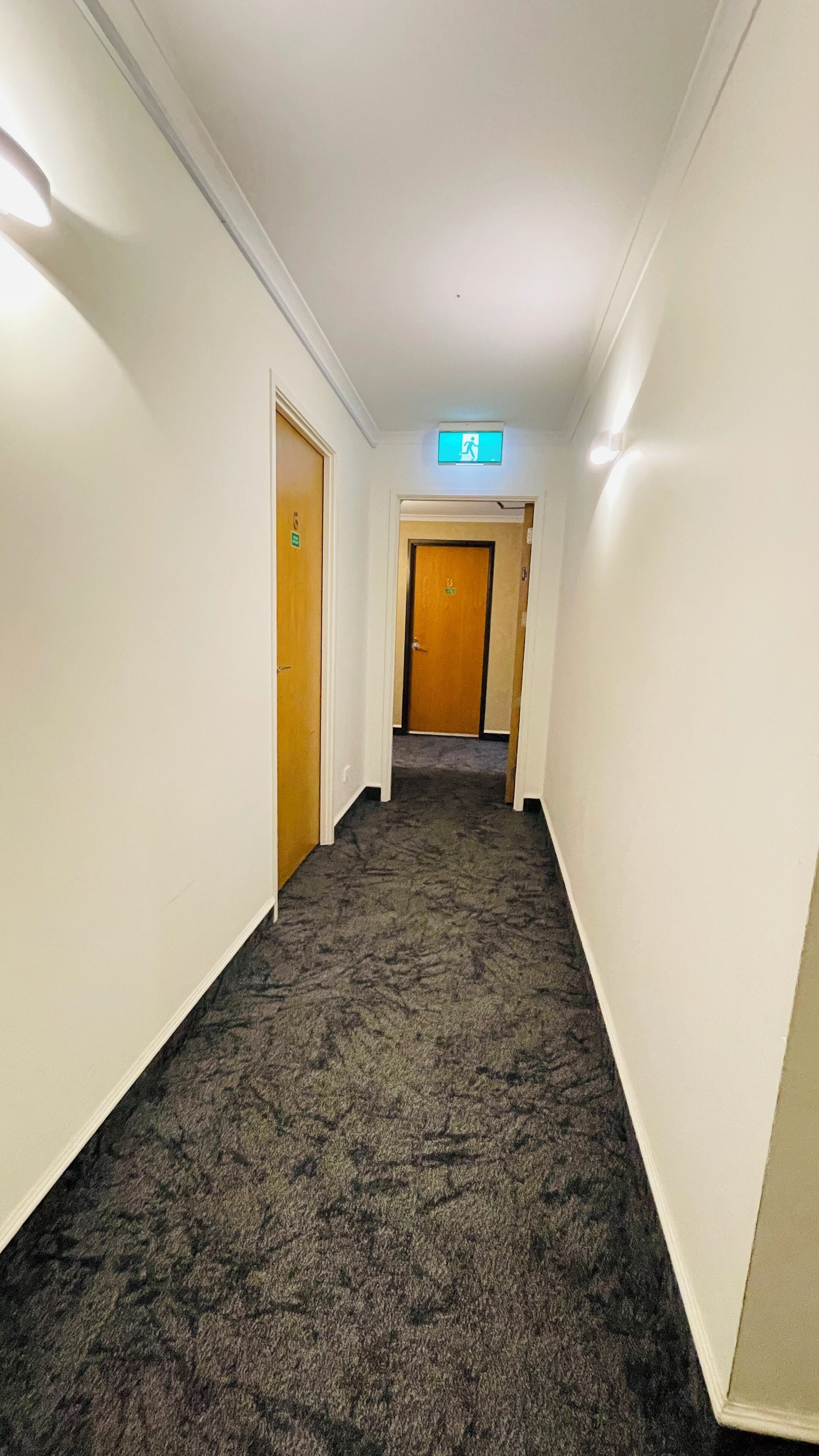 Pasillo alfombrado con paredes blancas, una puerta de madera al final y una señal de salida arriba.