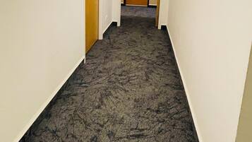 Pasillo alfombrado con paredes blancas, una puerta de madera al final y una señal de salida arriba.