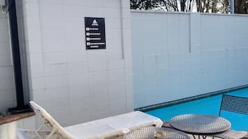 A poolside area with lounge chairs and tables, a sign prohibiting diving, running, bombing, unattended children, and a tiled wall.