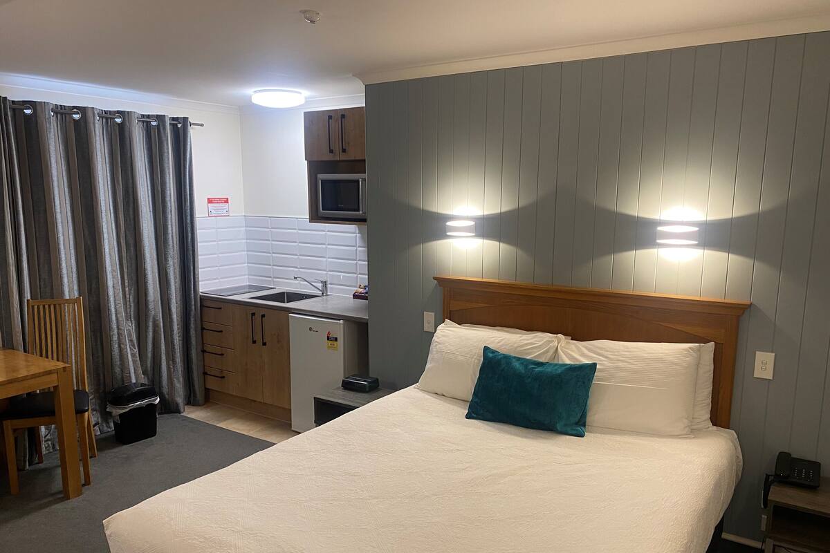 Una habitación de hotel compacta con una cama individual, un pequeño rincón americano, una silla de madera y una mesa de comedor.