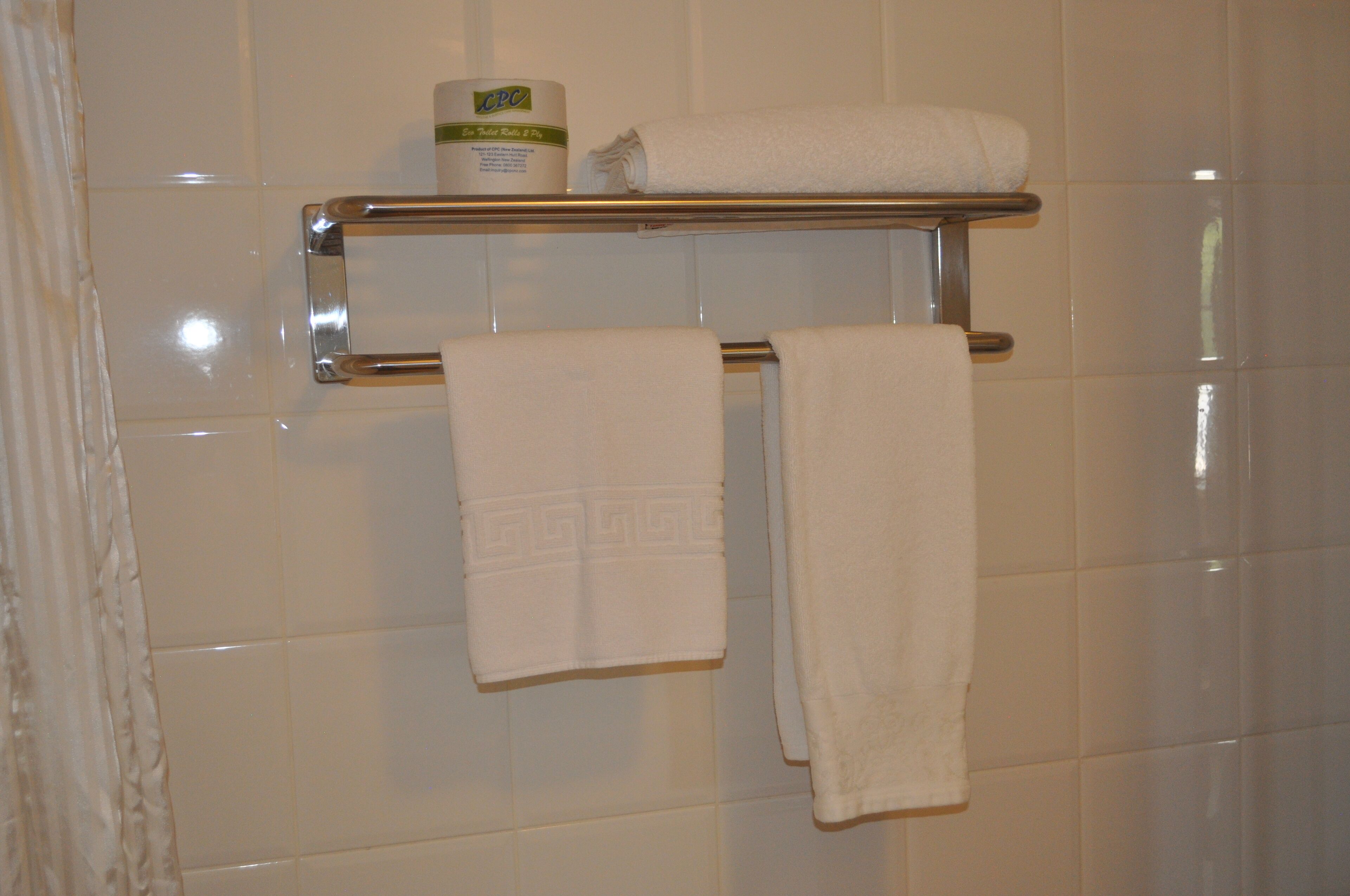 A bathroom with white tiled walls, a chrome towel rack, and a shelf holding a bottle of lotion and a rolled-up towel.