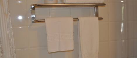 A bathroom with white tiled walls, a chrome towel rack, and a shelf holding a bottle of lotion and a rolled-up towel.