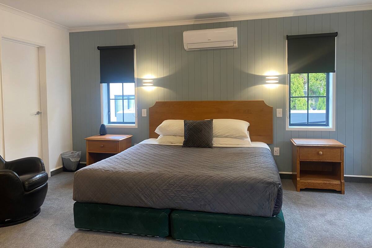 Un dormitorio con una cama, dos mesitas de noche, una mesita auxiliar, una silla, una ventana y un equipo de aire acondicionado.