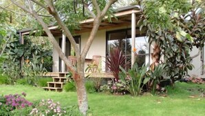 A house with a covered porch, surrounded by lush greenery and a well-maintained lawn. - Wharepuke Subtropical Accommodation (Kerikeri)