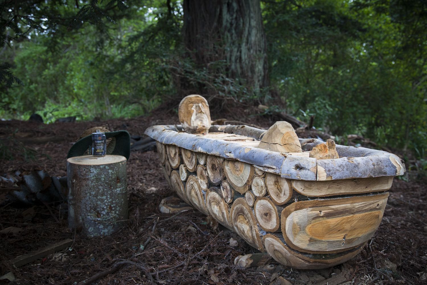 A rustic wooden boat with a log pattern design, placed on a forest floor with a tree trunk in the background.