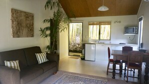 A compact living space with a sofa, dining table, chairs, a kitchen area, and a large window with a view of bamboo outside. - Wharepuke Subtropical Accommodation (Kerikeri)