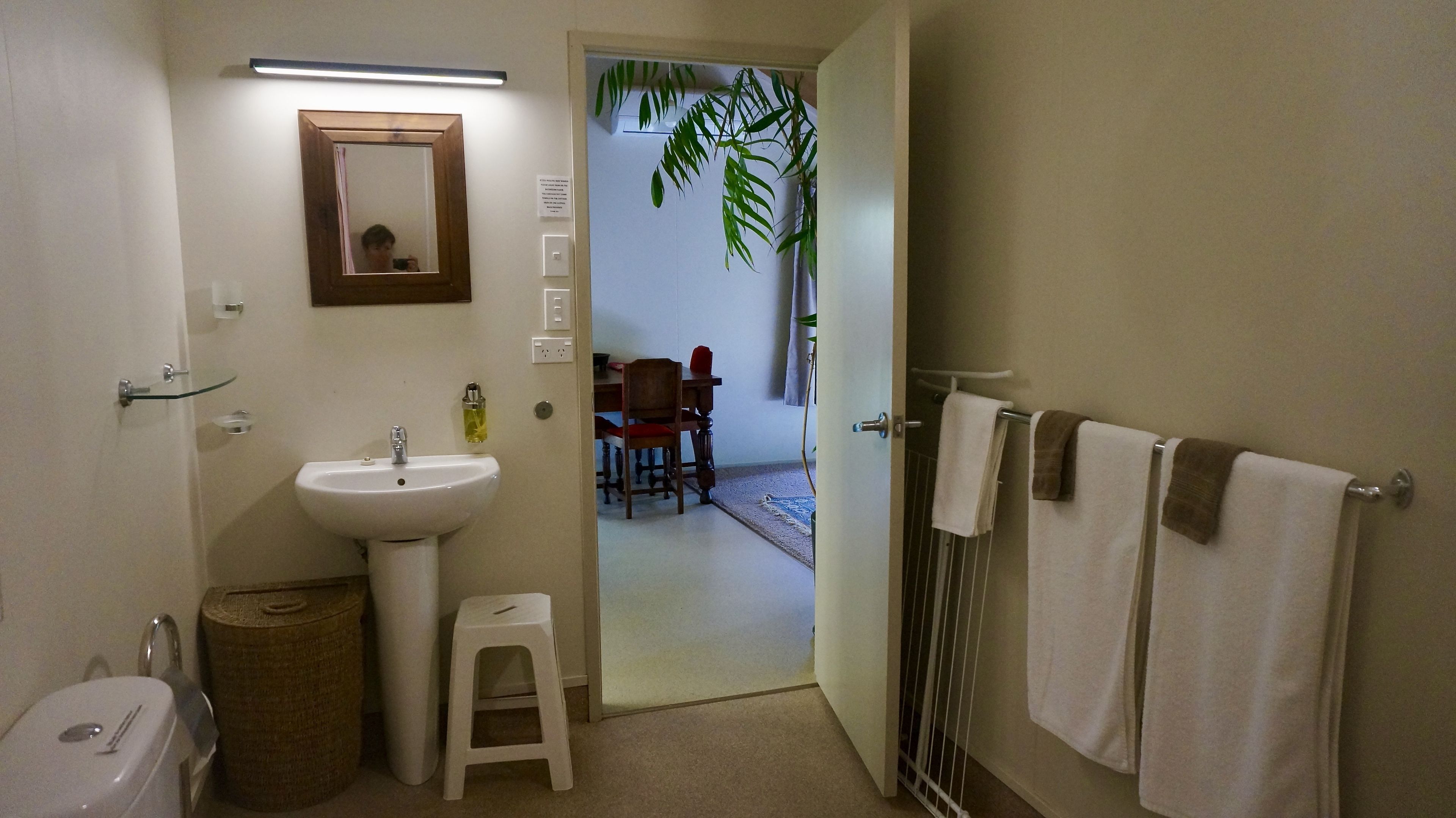 A bathroom with a sink, mirror, and towel rack. There is a small stool and a trash bin. The bathroom leads to a dining area with a table and chairs.