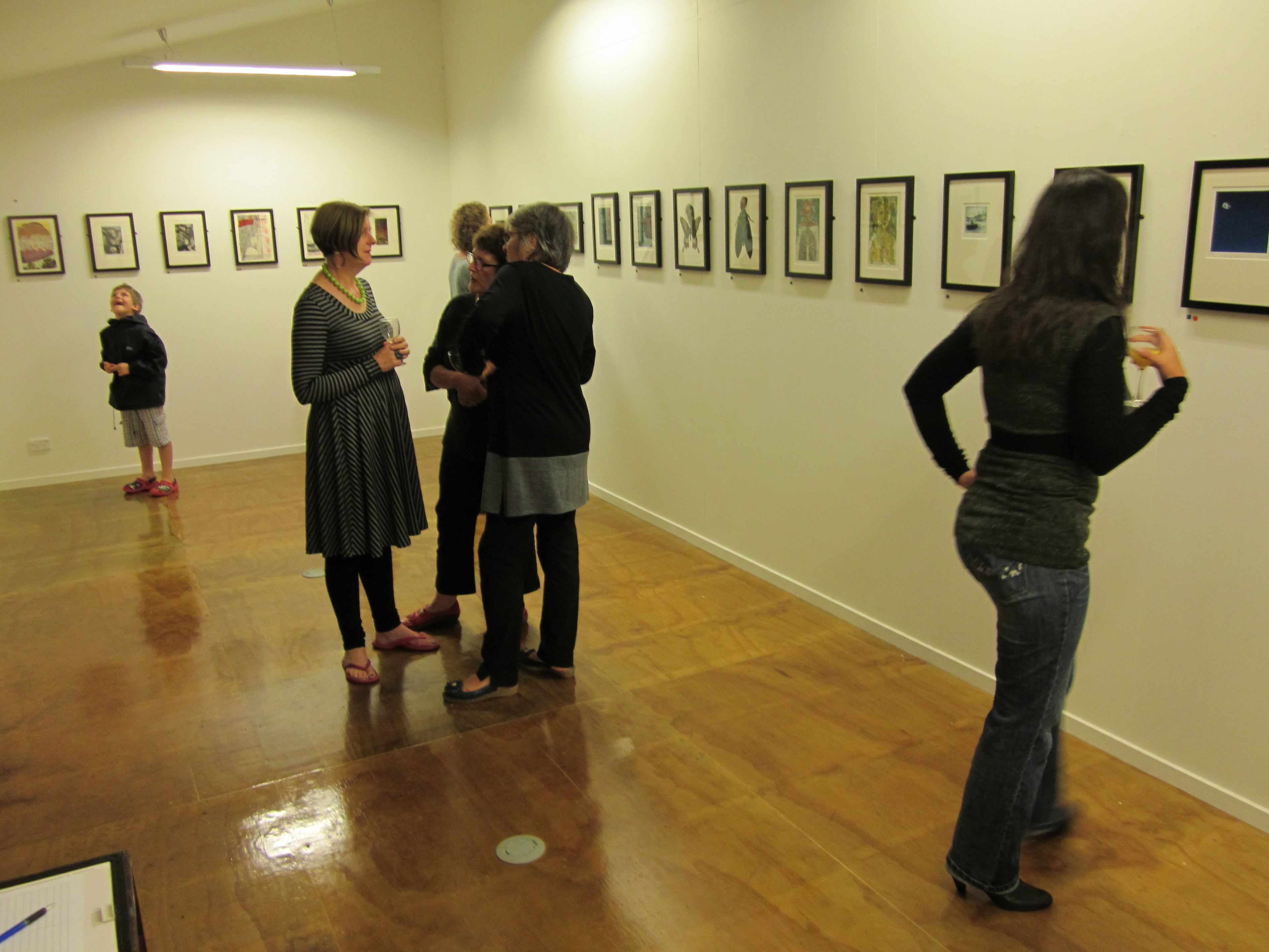 A gallery with framed artworks on the wall, wooden floors, and people viewing the art.