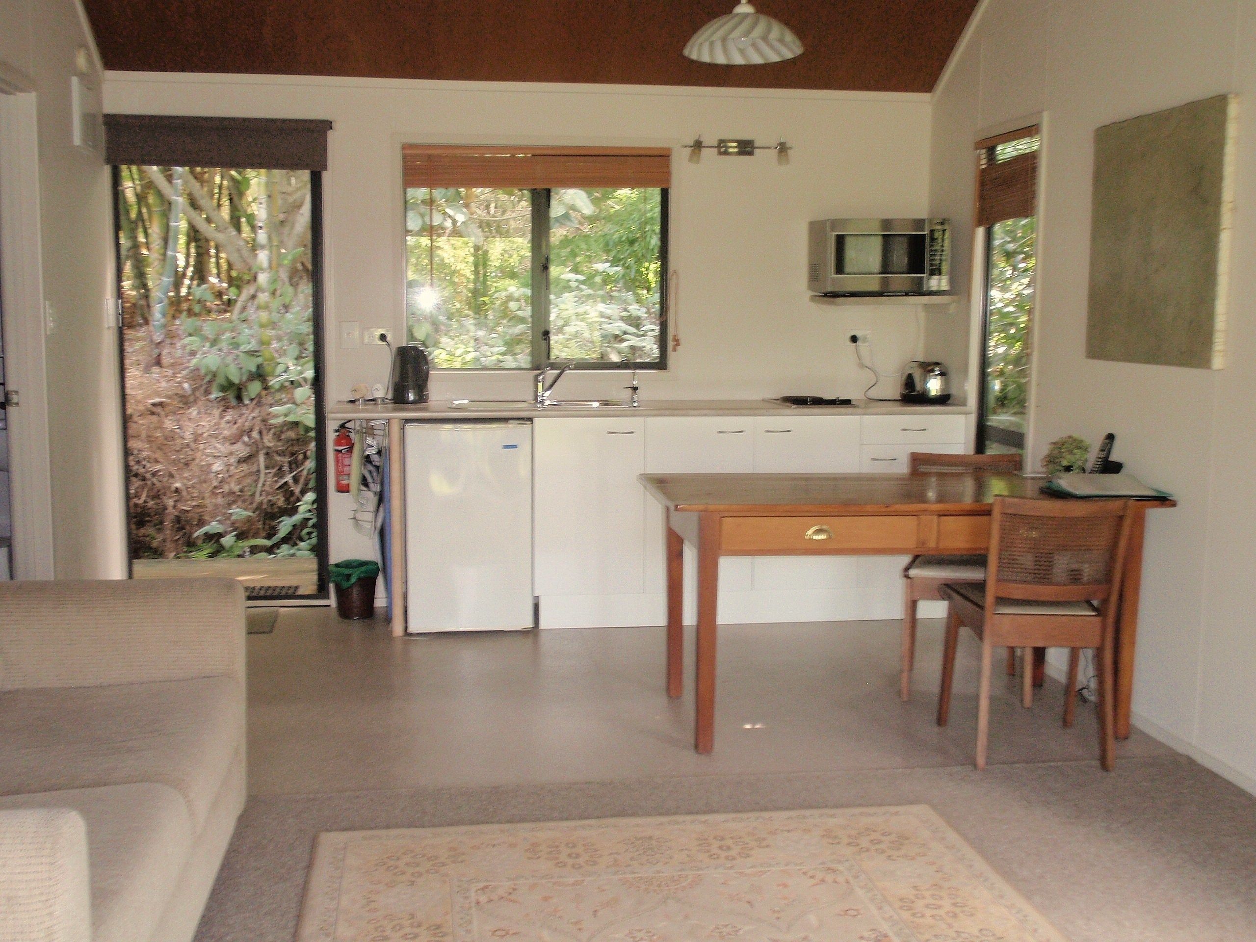 A compact kitchen with a microwave, sink, and small refrigerator. There's a dining table with chairs and a window with a view of greenery.