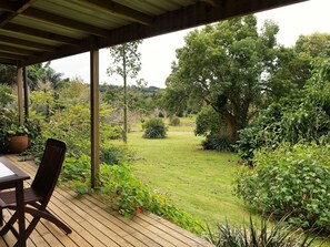 A covered patio with a wooden table and chair, overlooking a lush garden with various plants and trees. - Wharepuke Subtropical Accommodation (Kerikeri)