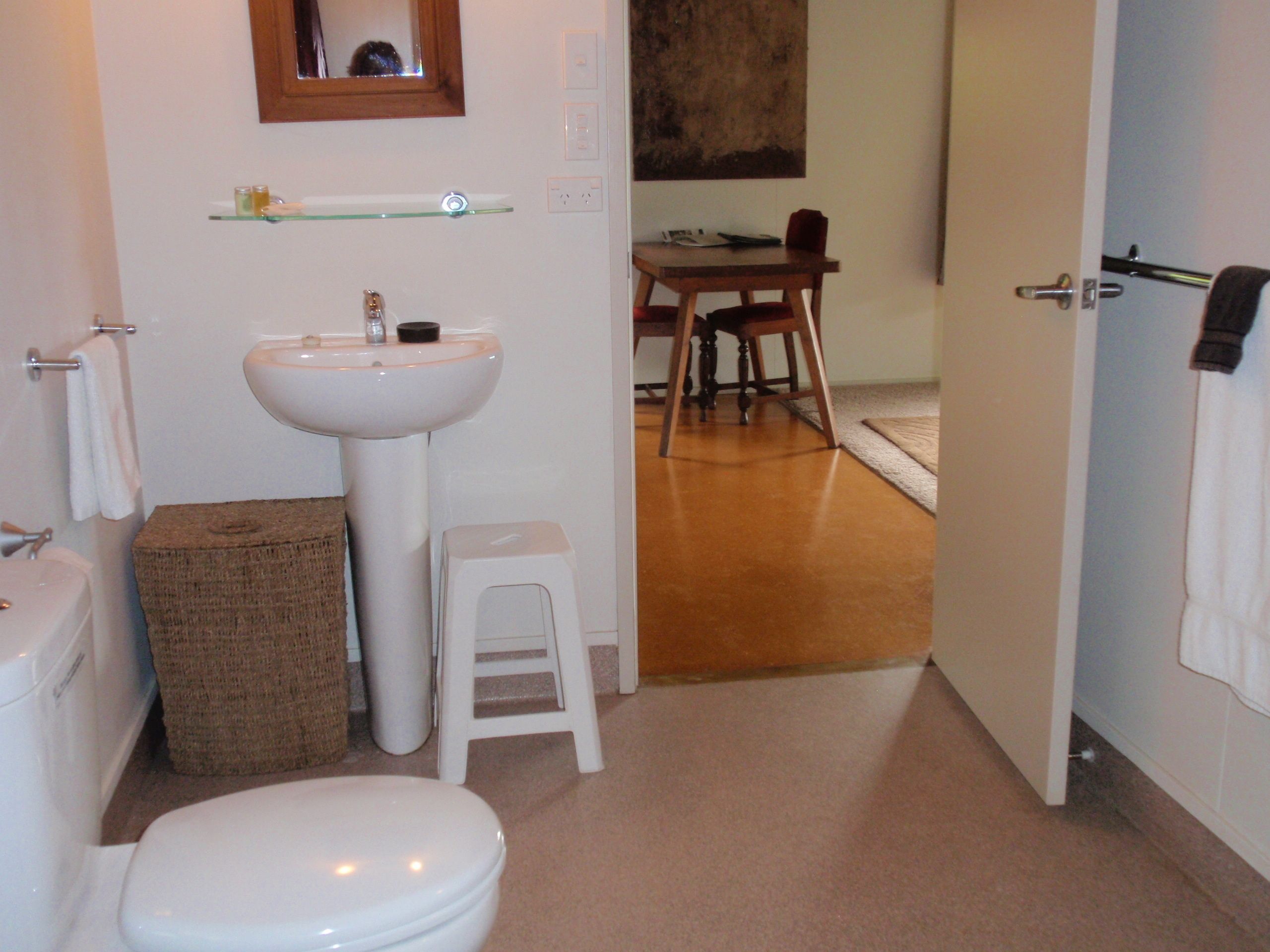 Ein Badezimmer mit Toilette, Waschbecken und Spiegel. Ein kleiner Hocker und ein Korb befinden sich in der Nähe des Waschbeckens.