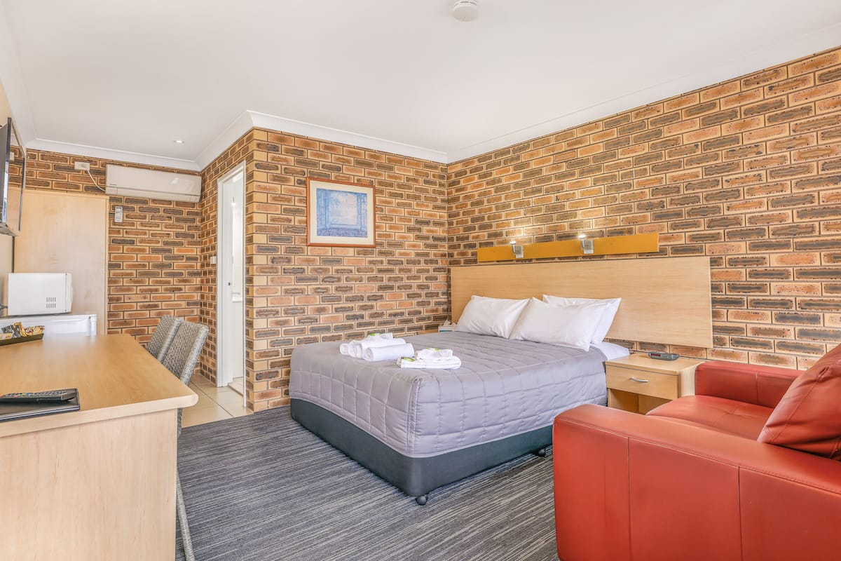 A hotel room with a brick wall, a bed with a grey quilt, a wooden desk, a red chair, and a small bedside table with a lamp.