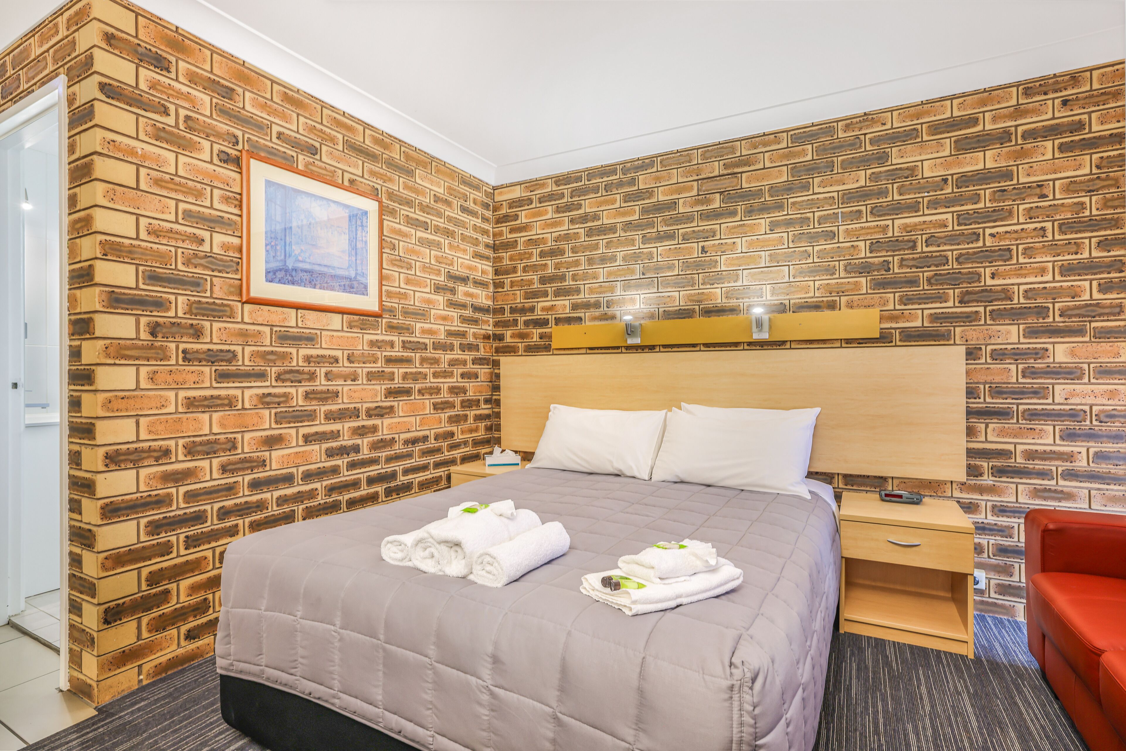 Ein Hotelzimmer mit einer Wand im Backstein-Look, einem Bett mit einer beige farbenen Bettdecke, zwei ordentlich gefalteten Handtüchern, einem Nachttisch aus Holz und einem roten Sessel.