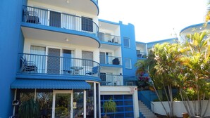 Un hotel de varios pisos con balcones azules, una entrada cubierta y un área ajardinada con plantas.