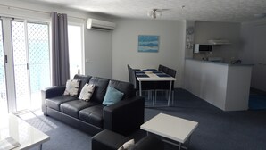 Una sala moderna con un sofá de cuero negro, una mesa de centro blanca y un comedor con una mesa y sillas blancas. Hay una zona de cocina con estufa y microondas.
