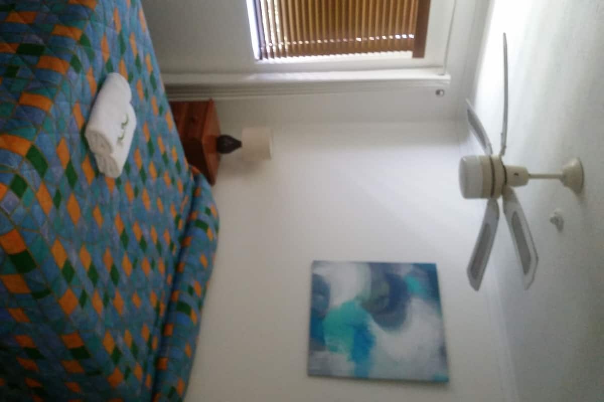 A room with a bed covered in a colorful quilt, a white towel on the bed, a ceiling fan, and a painting on the wall.