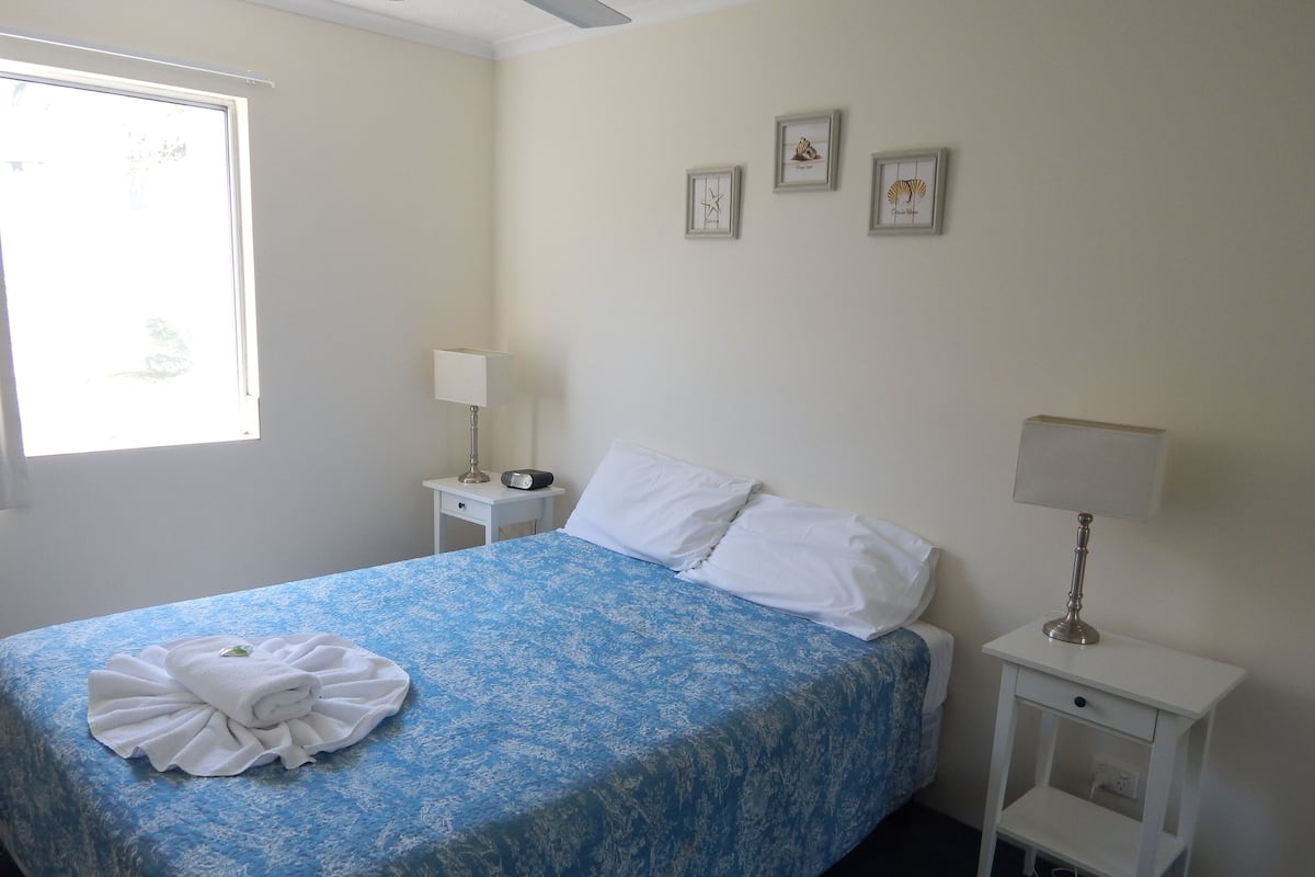 A neatly made bed with a blue floral bedspread, white pillows, and a folded white towel on the left side. There are three framed pictures on the wall, a bedside table with a lamp, and a window with curtains.
