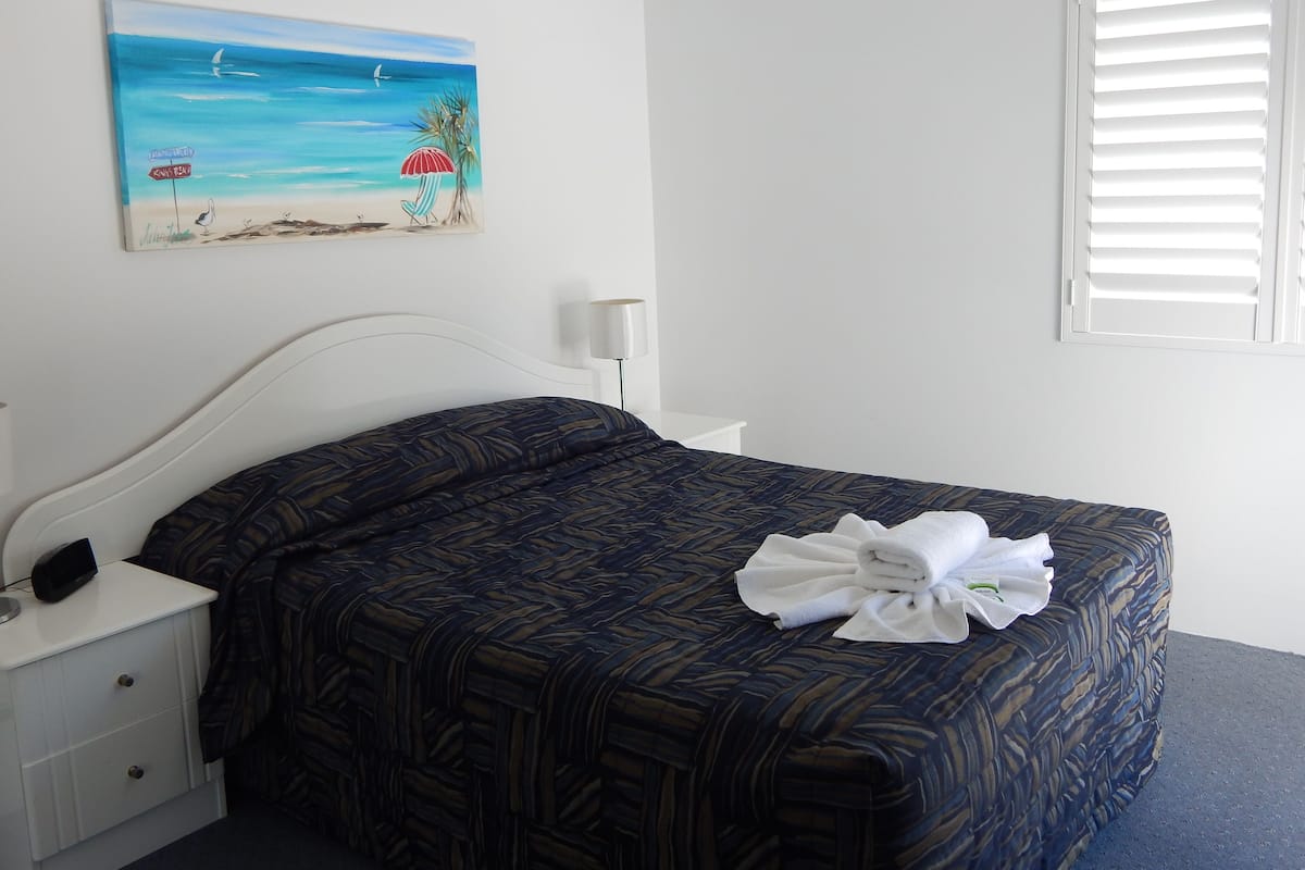 A neatly made bed with a patterned bedspread, a white bedside table with a lamp, and a painting of a beach scene on the wall.
