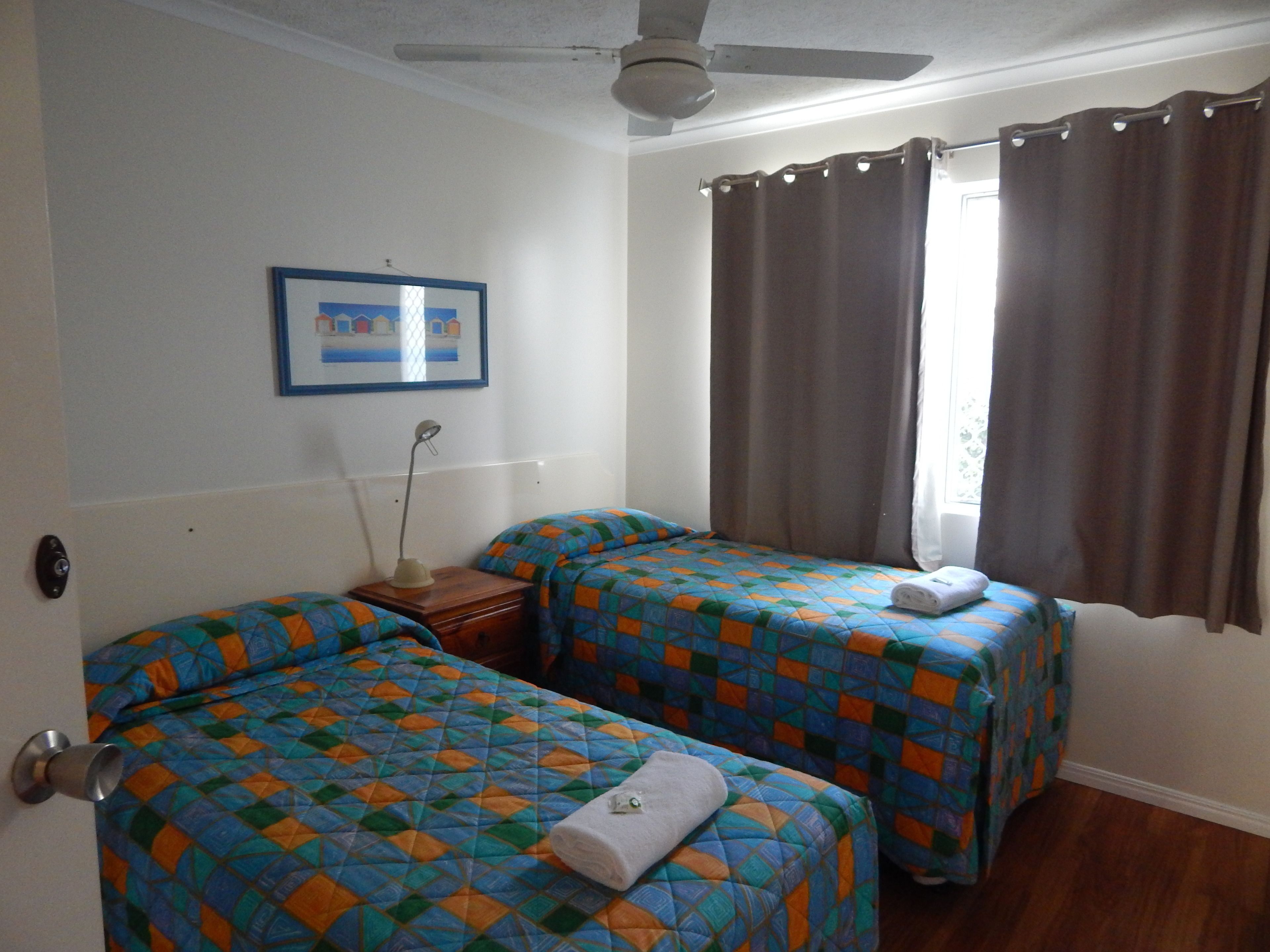 Une chambre avec deux lits, un ventilateur de plafond, un tableau encadré au mur et une fenêtre avec des rideaux.