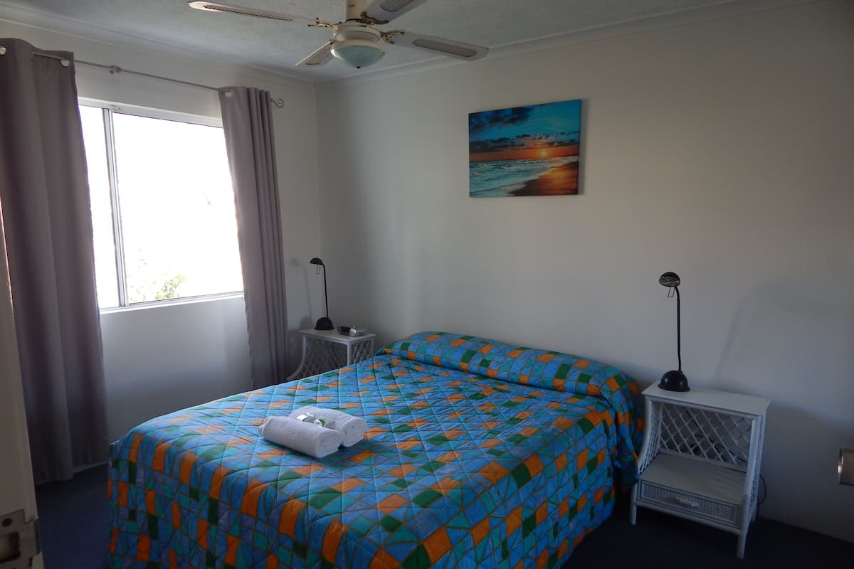 A hotel room with a bed, two bedside tables, a ceiling fan, a painting of a sunset, and a window with curtains.
