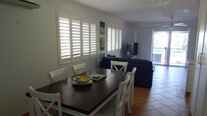 Un comedor con una mesa de madera oscura, sillas blancas y un sofá azul. Hay ventanas con persianas, un ventilador de techo y un televisor sobre un mueble.