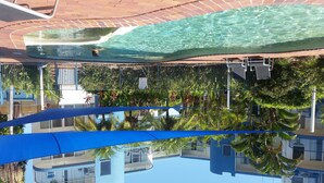 Zona de piscina con agua cristalina, rodeada de palmeras y toldos azules.