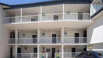 A multi-story building with a flat roof, balconies on each floor, and a parking area in front.