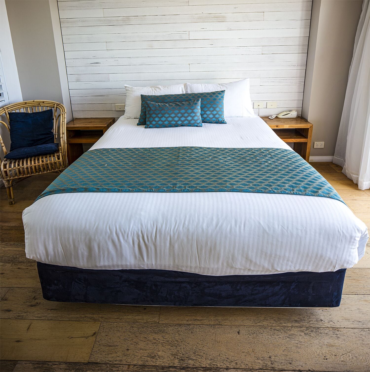 Un lit bien fait, recouvert d’une couette blanche et turquoise, avec deux oreillers et une table de chevet en bois sur laquelle se trouve un téléphone.