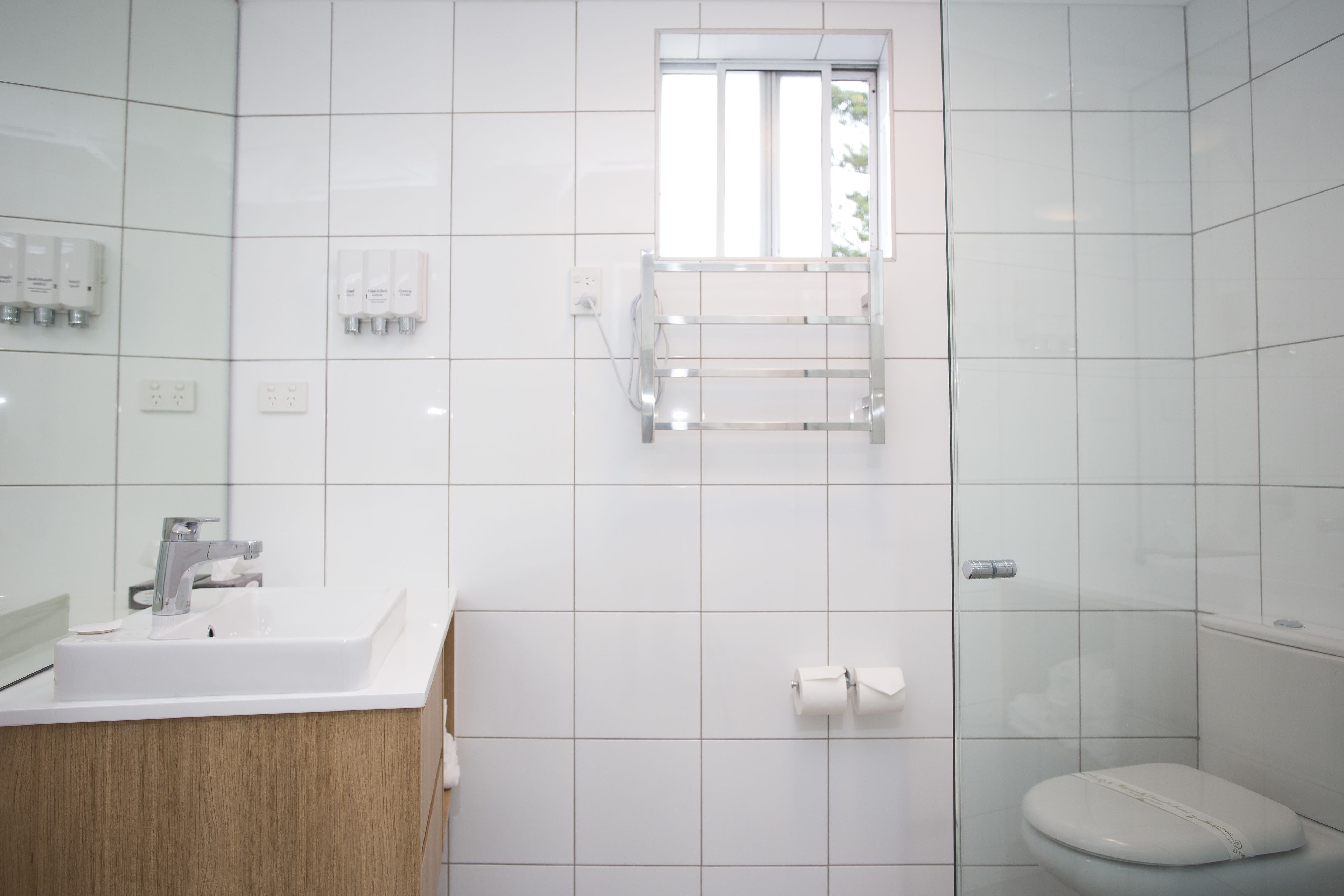 A modern bathroom with a white sink, mirror, and toilet. There is a window above the toilet, and a towel rack mounted on the wall.