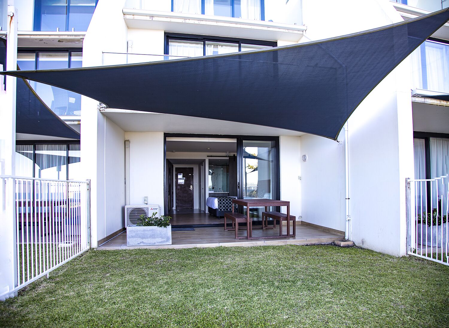 Een modern gebouw met een overdekte patio, een tafel, stoelen, een kleine tuin en een witte schutting.
