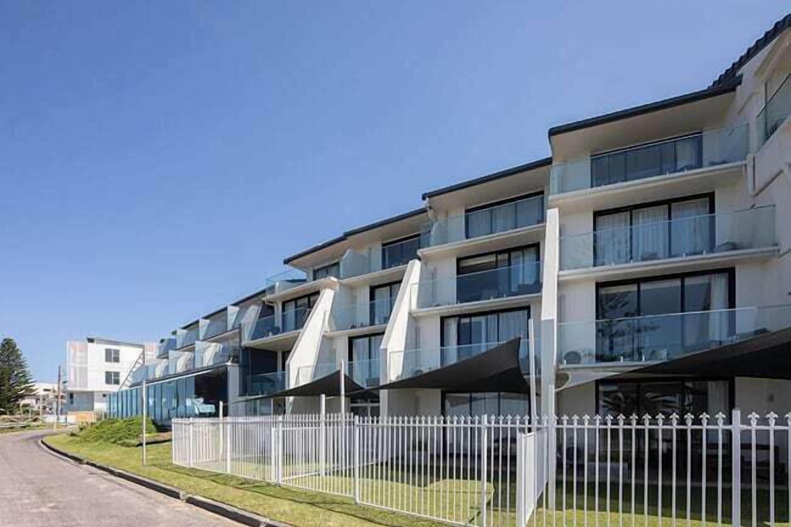 A modern multi-story residential building with balconies, a white fence, and a clear sky.