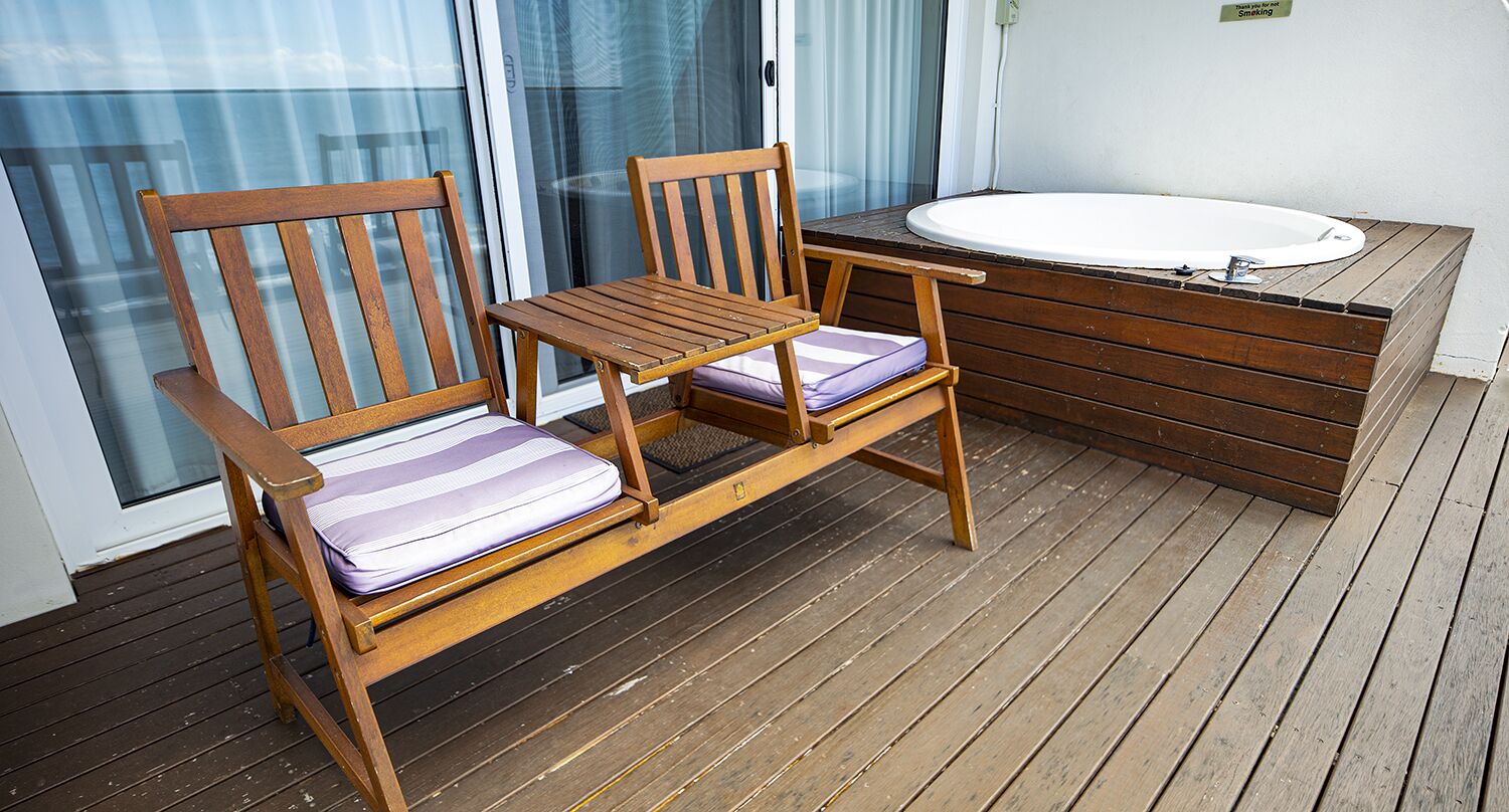 A wooden deck with two wooden chairs and a small table, a white bathtub, and a glass door leading to a balcony.