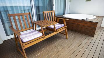 A wooden deck with two wooden chairs and a small table, a white bathtub, and a glass door leading to a balcony.