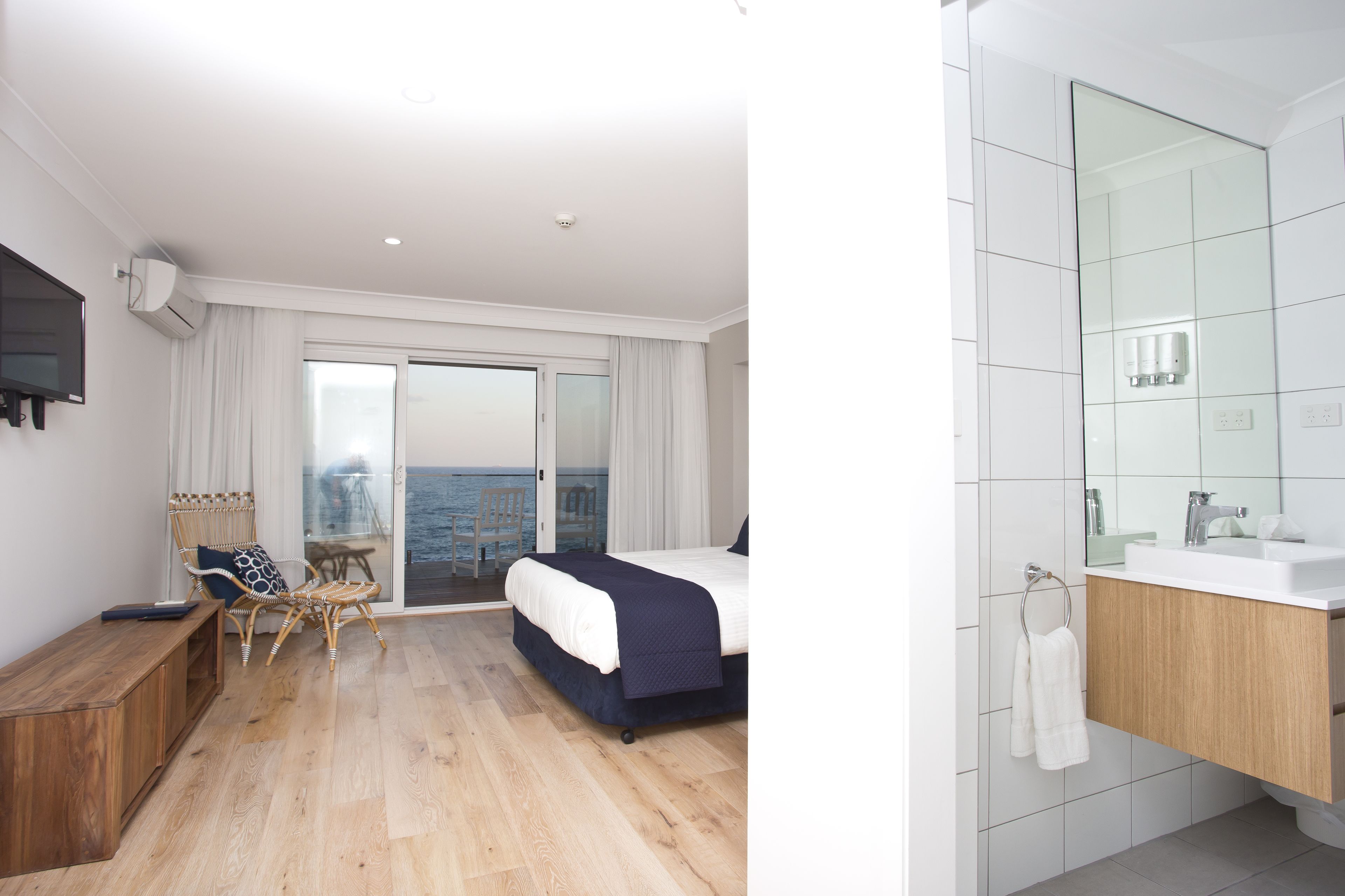 Una habitación de hotel moderna con cama, un banco de madera, una silla de mimbre, un balcón con vistas al mar y un baño con un espejo grande y lavamanos.