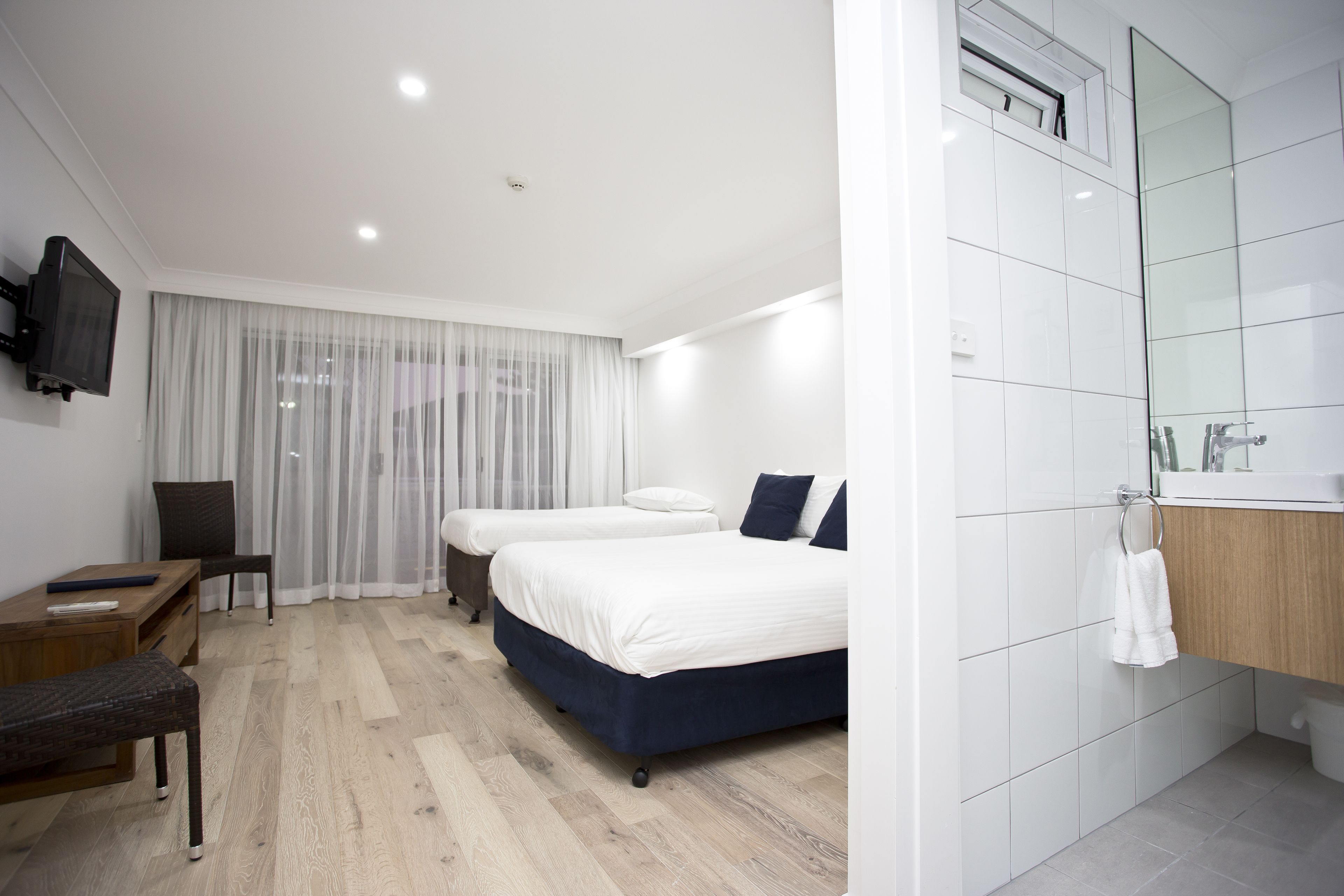 A modern hotel room with a wooden floor, two beds, a TV, a desk, and a bathroom with a mirror and sink.
