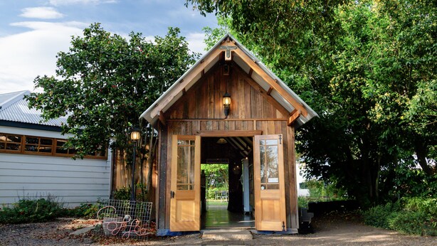 A rustic wooden structure with a sloped roof and a cross on top, likely a chapel or small church, surrounded by lush greenery.