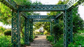 A stone pathway leading through a green metal archway surrounded by lush greenery.