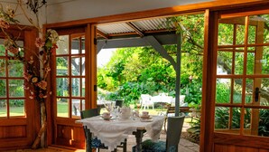 A dining area with a table set for tea, surrounded by chairs, and a view of a garden through large windows.