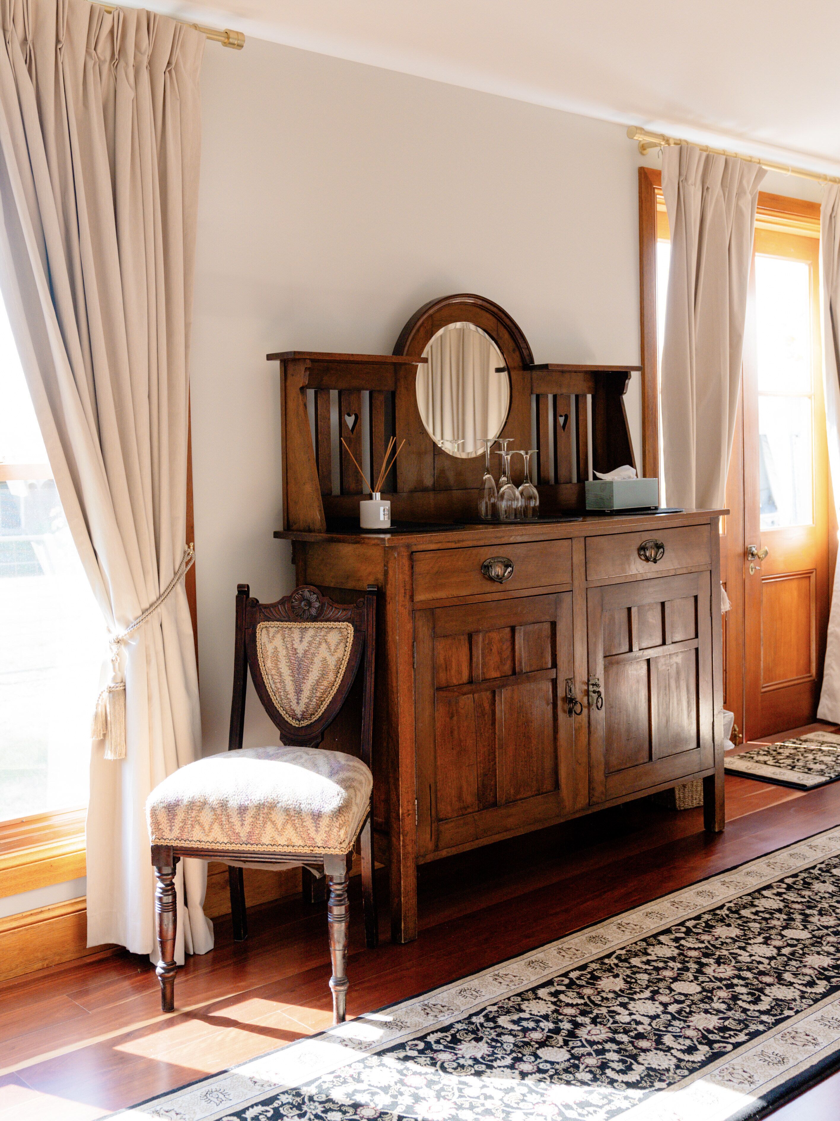 Un aparador de madera con espejo, una silla y un tapete estampado en una habitación con suelo de madera y cortinas.