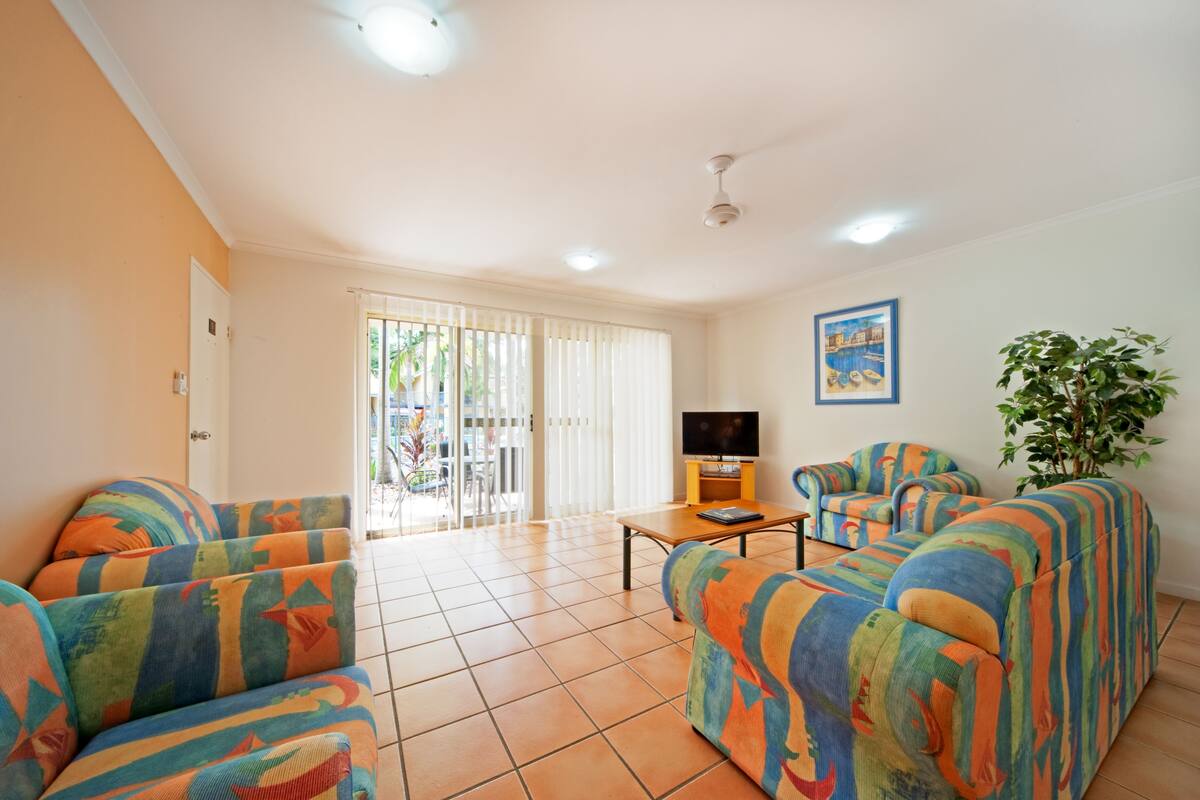 Una sala de estar con sofás coloridos, un televisor sobre una mesa de madera y un ventanal con persianas.