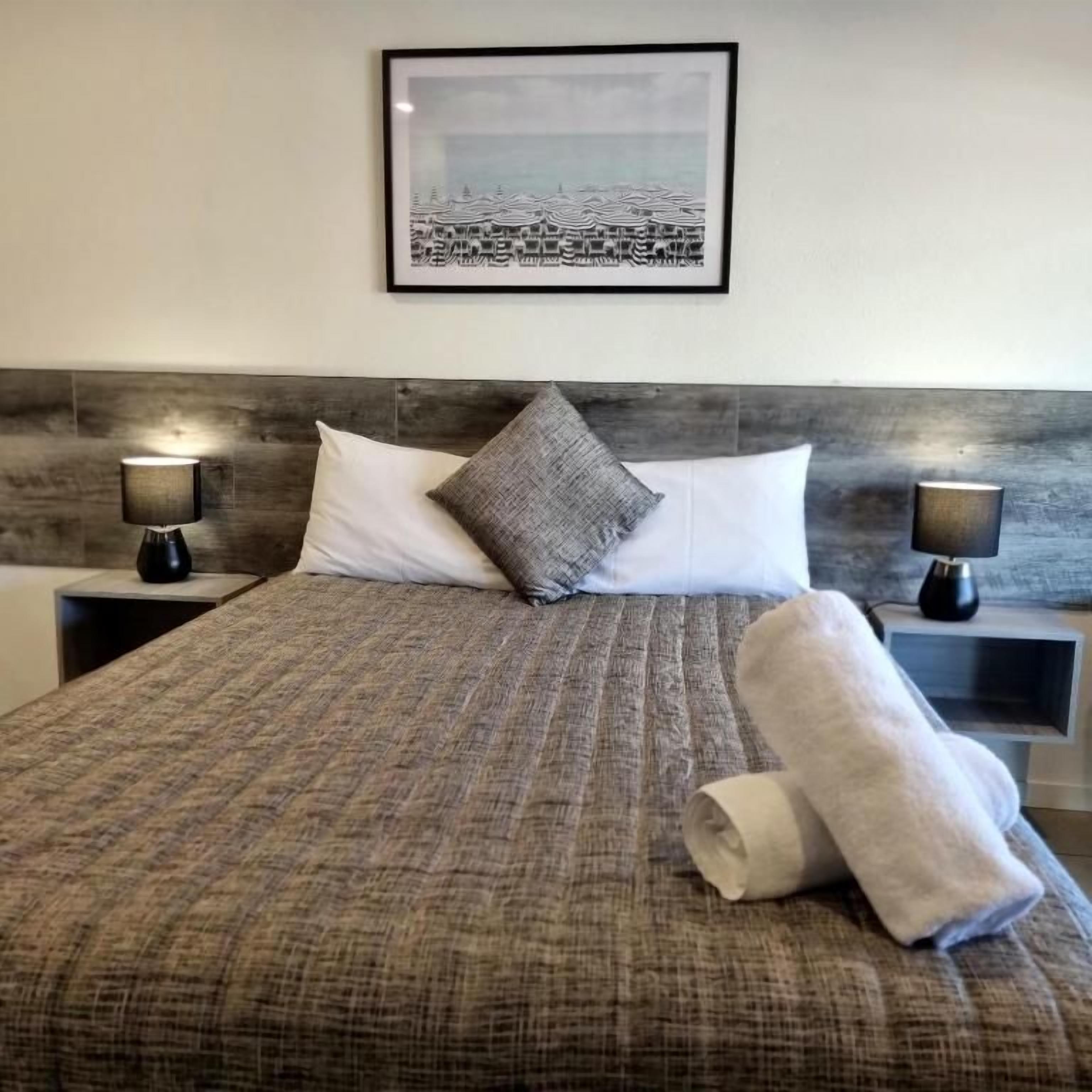 A neatly made bed with a patterned bedspread, two pillows, and a rolled towel on the side. There are two bedside lamps on either side of the bed. A framed picture hangs on the wall above the bed.