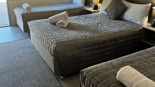 Habitación de hotel con una cama grande, un sofá más pequeño, una mesita de noche con lámpara y una ventana con persianas.