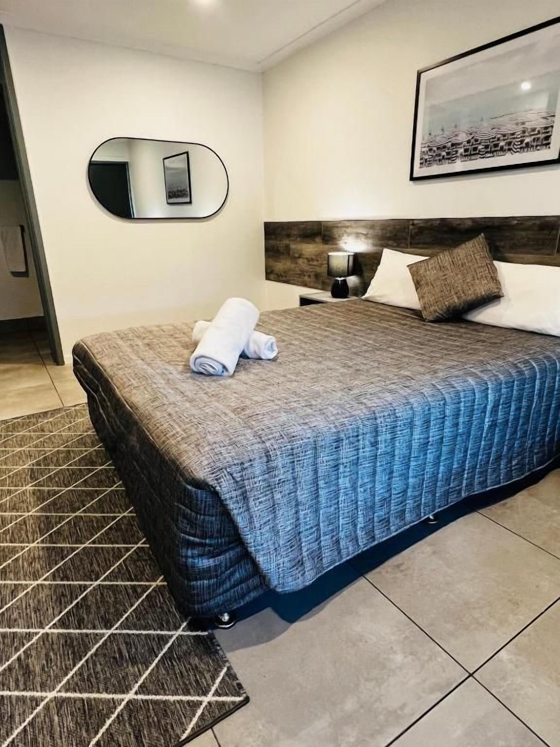 Habitació d'hotel amb un llit, un capçal, una petita taula de nit, una làmpada, un mirall i un quadre amb marc a la paret.