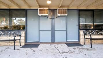 Entrada de un edificio con dos unidades de aire acondicionado, un conjunto de puertas dobles y dos bancos afuera.