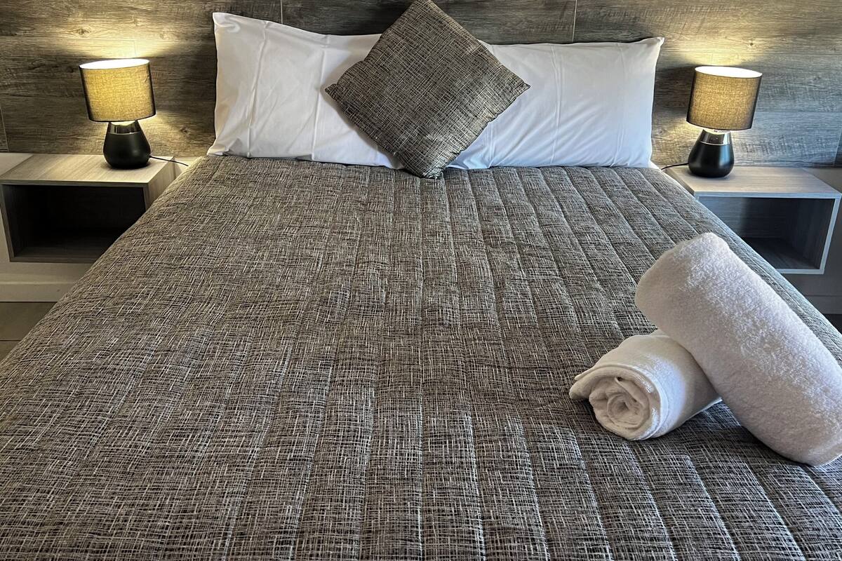 Una cama bien tendida con un edredón gris texturizado, dos almohadas blancas, una almohada gris y una toalla blanca enrollada. Hay dos lámparas de noche a ambos lados de la cama.