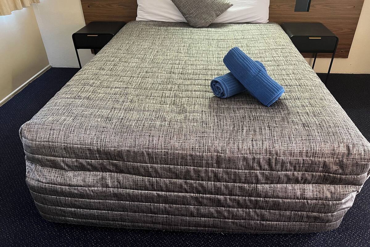 Una cama bien hecha con un edredón gris y blanco estampado, dos almohadas y toallas azules enrolladas sobre una alfombra oscura con vetas.