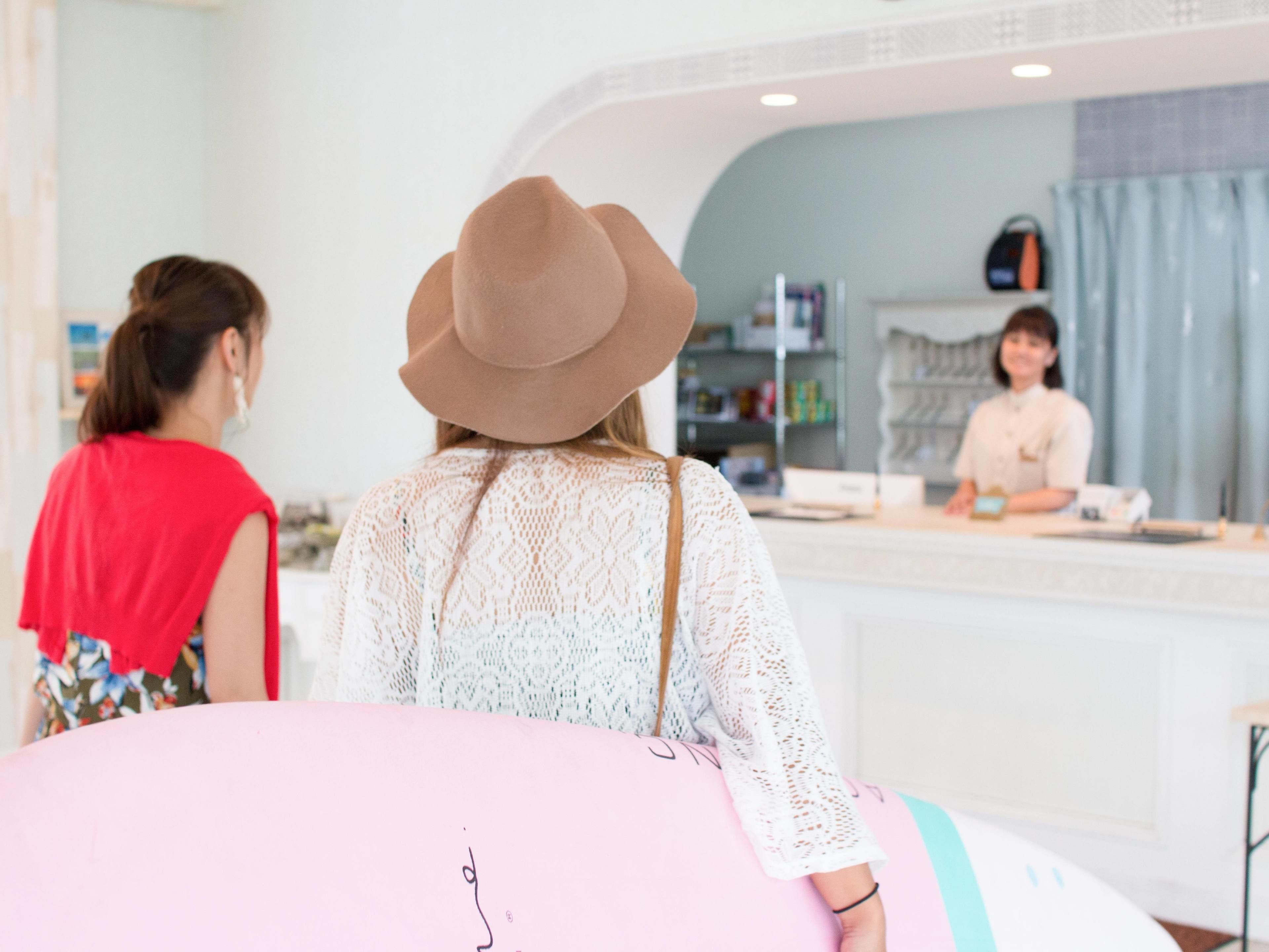 A person with a pink surfboard and a hat at a counter in a room with a white wall and a blue curtain.