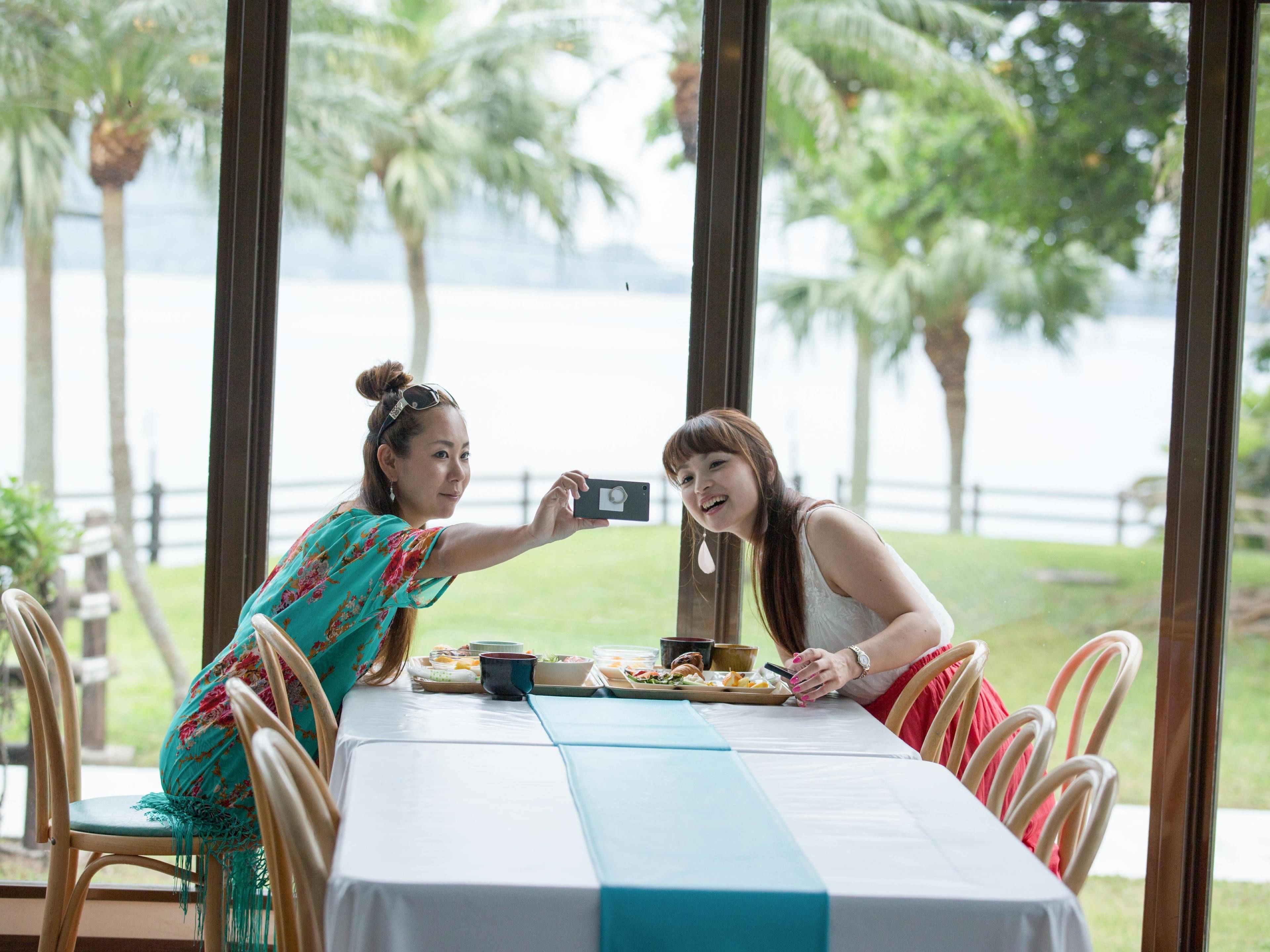 Deux personnes prenant un selfie à une table, avec en arrière-plan des palmiers et l’eau.