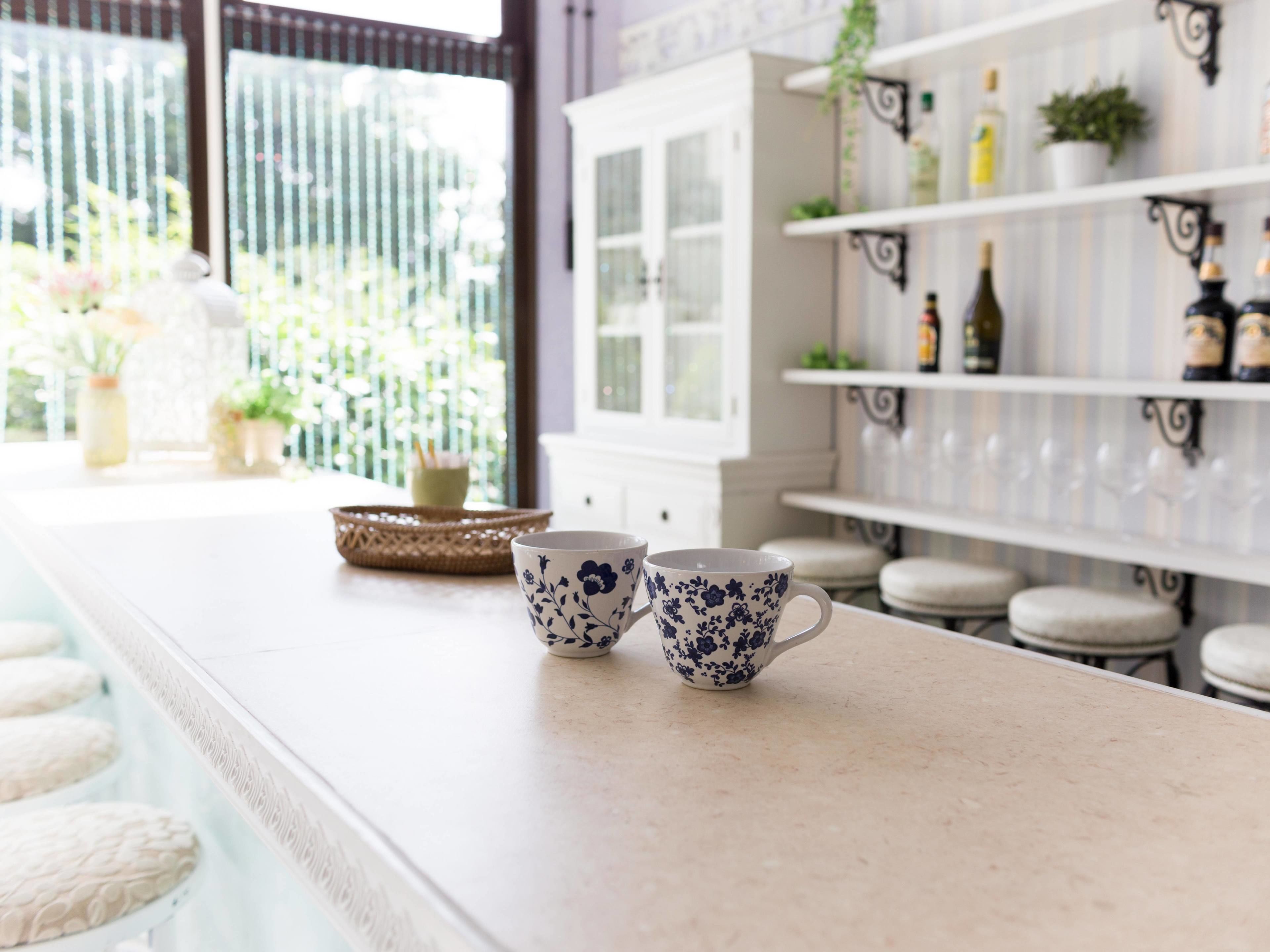 A kitchen counter with two floral-patterned cups, a wicker basket, and a white cabinet with glass doors.